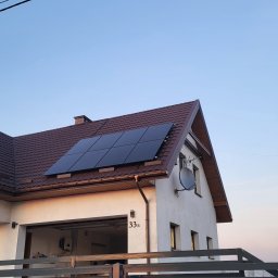 Zielony Program Ekologiczny sp. z o.o. - Dom jednorodzinny z brązowym dachem, na którym zamontowano czarne panele słoneczne. W tle błękitne niebo, a na pierwszym planie nowoczesne, szare ogrodzenie.