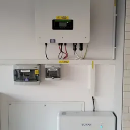 Biała instalacja fotowoltaiczna Sofar na ścianie, widoczne: inwerter, rozdzielnice i akumulator energii, kable i rury prowadzone wzdłuż ściany.