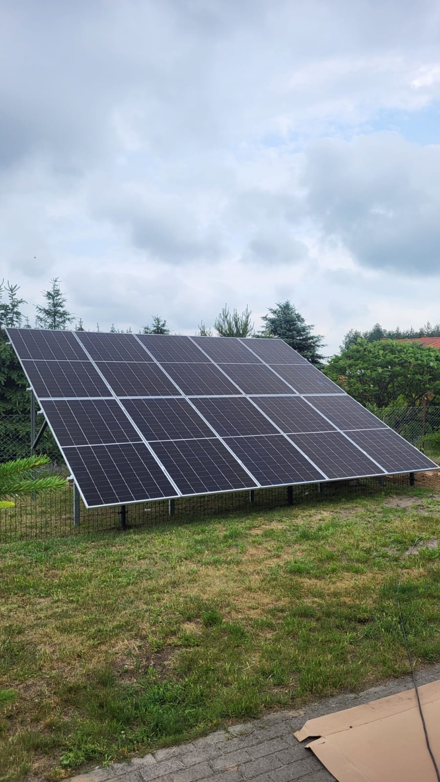 Zestaw paneli słonecznych na metalowej konstrukcji, zainstalowany na trawniku w ogrodzie, widok z dołu, pochmurne niebo w tle.