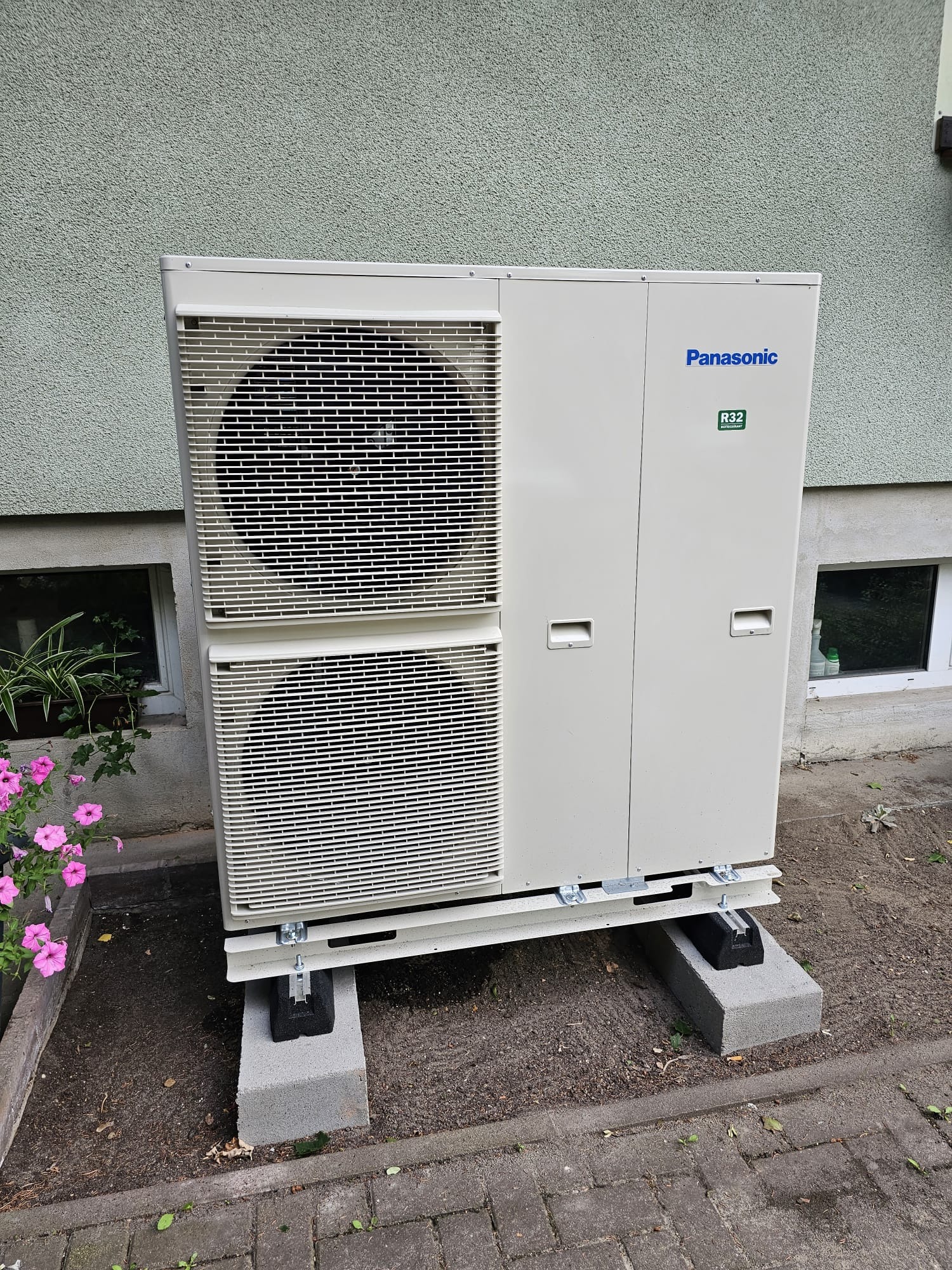 Zewnętrzna jednostka klimatyzacyjna Panasonic R32 na betonowych podstawach, z kwiatami w tle, ustawiona przy ścianie budynku.