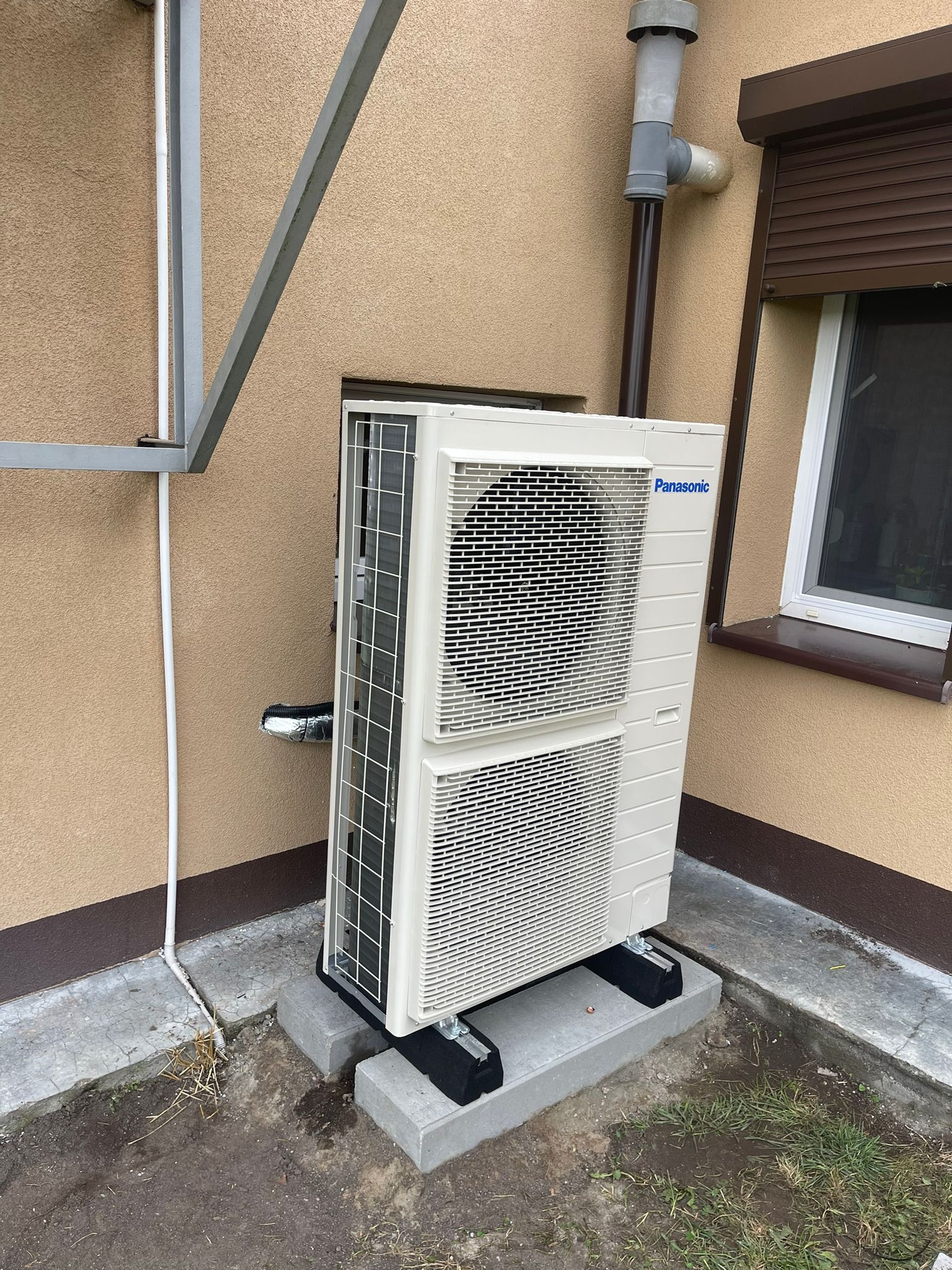 Zewnętrzna jednostka klimatyzacyjna Panasonic, zamontowana na betonowych podstawach przy ścianie budynku z brązową stolarką okienną i rynną odpływową, widoczne elementy instalacji.