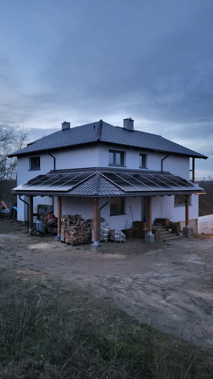 Dwukondygnacyjny dom jednorodzinny z panelami słonecznymi zamontowanymi na zadaszeniu tarasu; na podwórzu widoczne materiały budowlane i betoniarka.