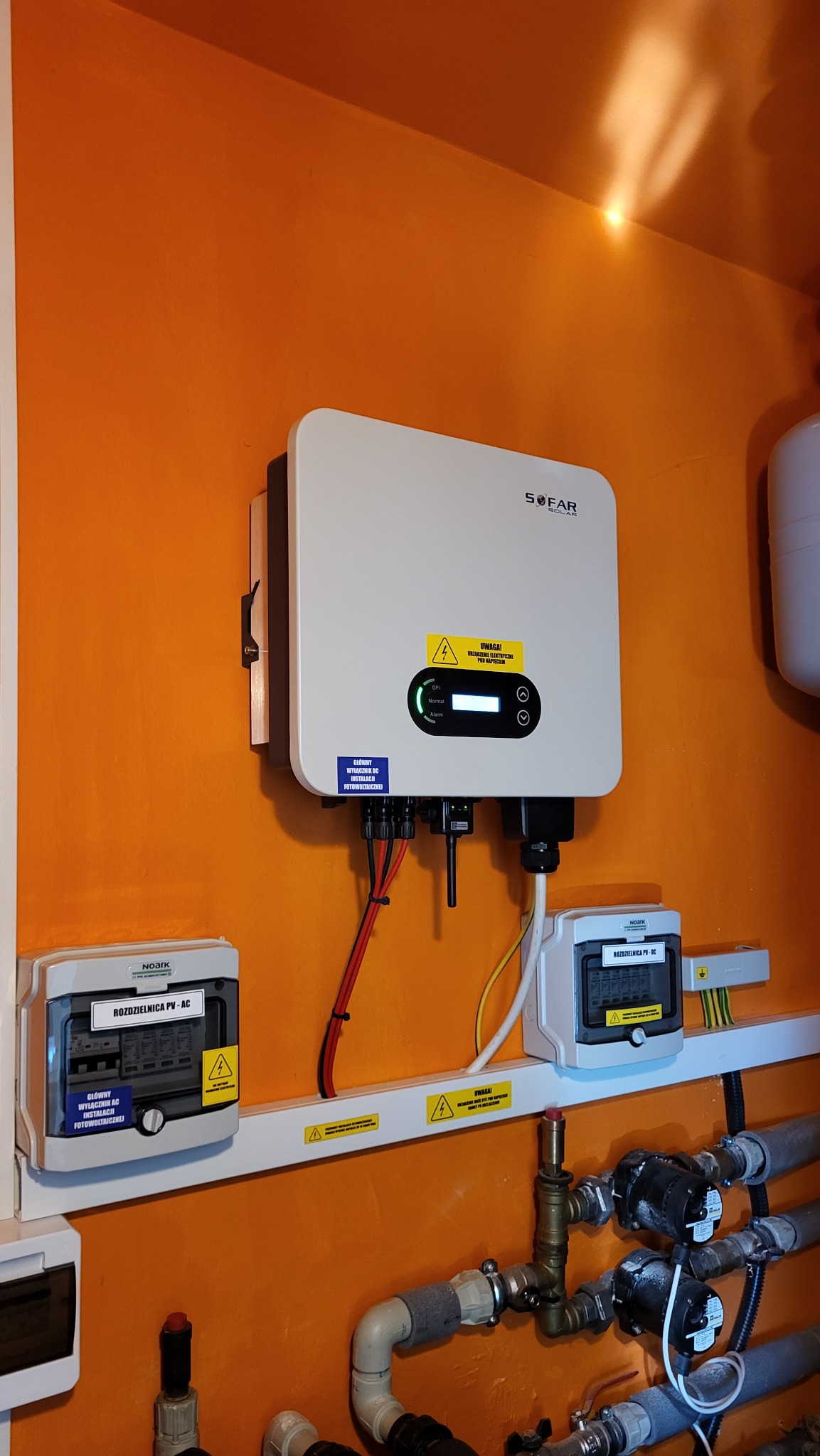 Biały falownik SOFAR zamontowany na pomarańczowej ścianie, pod nim skrzynki rozdzielcze PV-AC i PV-DC, widoczne rury instalacji.