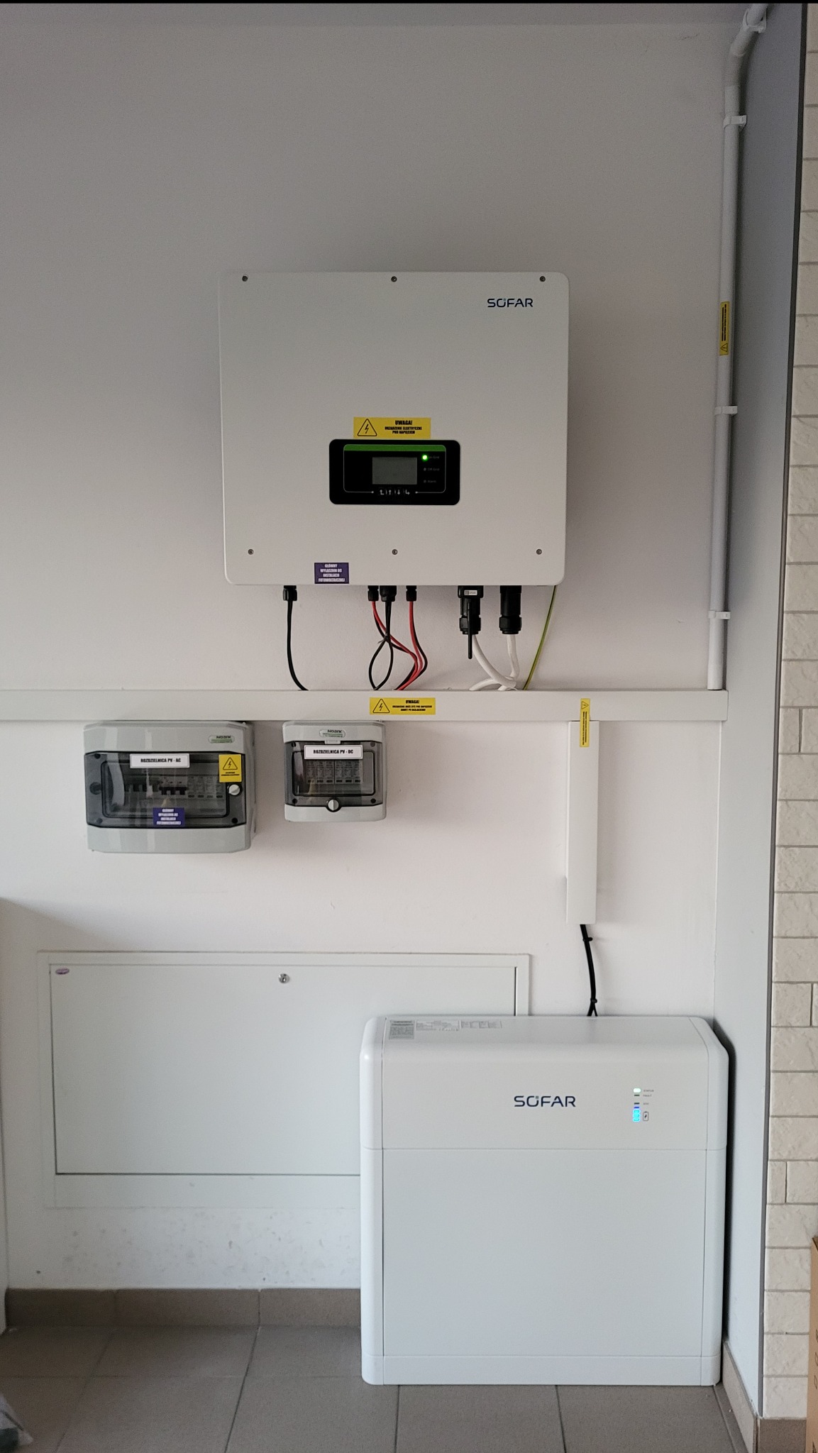 Biała instalacja fotowoltaiczna Sofar na ścianie, widoczne: inwerter, rozdzielnice i akumulator energii, kable i rury prowadzone wzdłuż ściany.