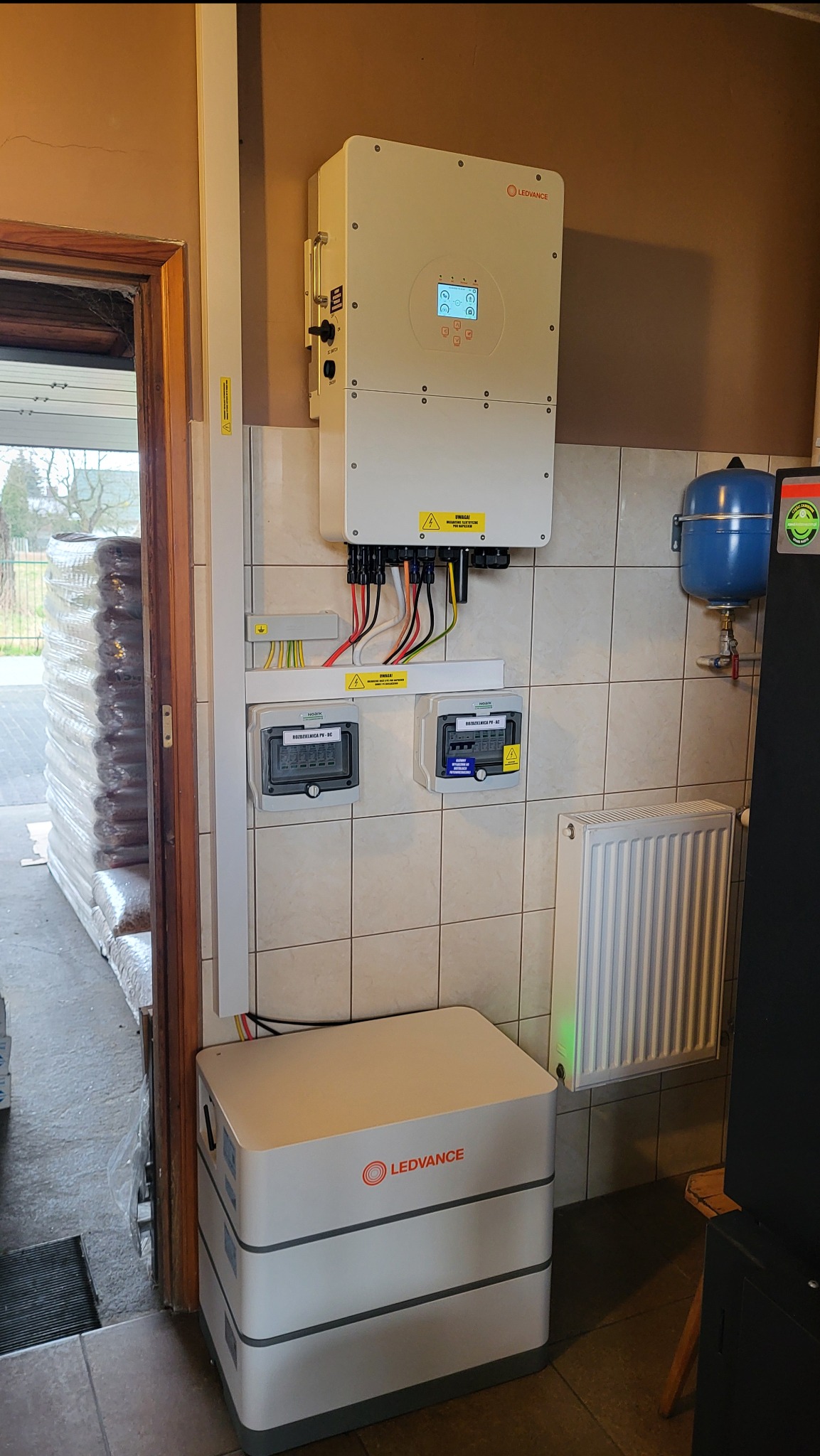 Instalacja fotowoltaiczna w pomieszczeniu gospodarczym: inwerter Ledvance z wyświetlaczem, akumulator, dwa liczniki energii elektrycznej na kafelkowanej ścianie, obok grzejnik i niebieski zbiornik...