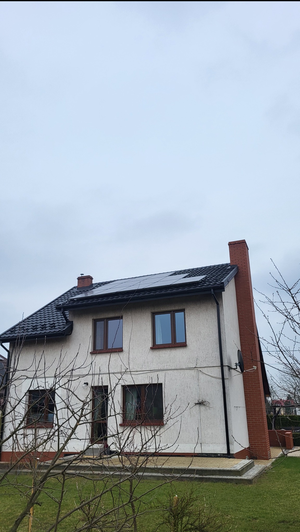 Dwupiętrowy dom z ciemną dachówką i panelami słonecznymi na dachu, widoczny komin z czerwonej cegły i drzewo na pierwszym planie, pochmurne niebo.