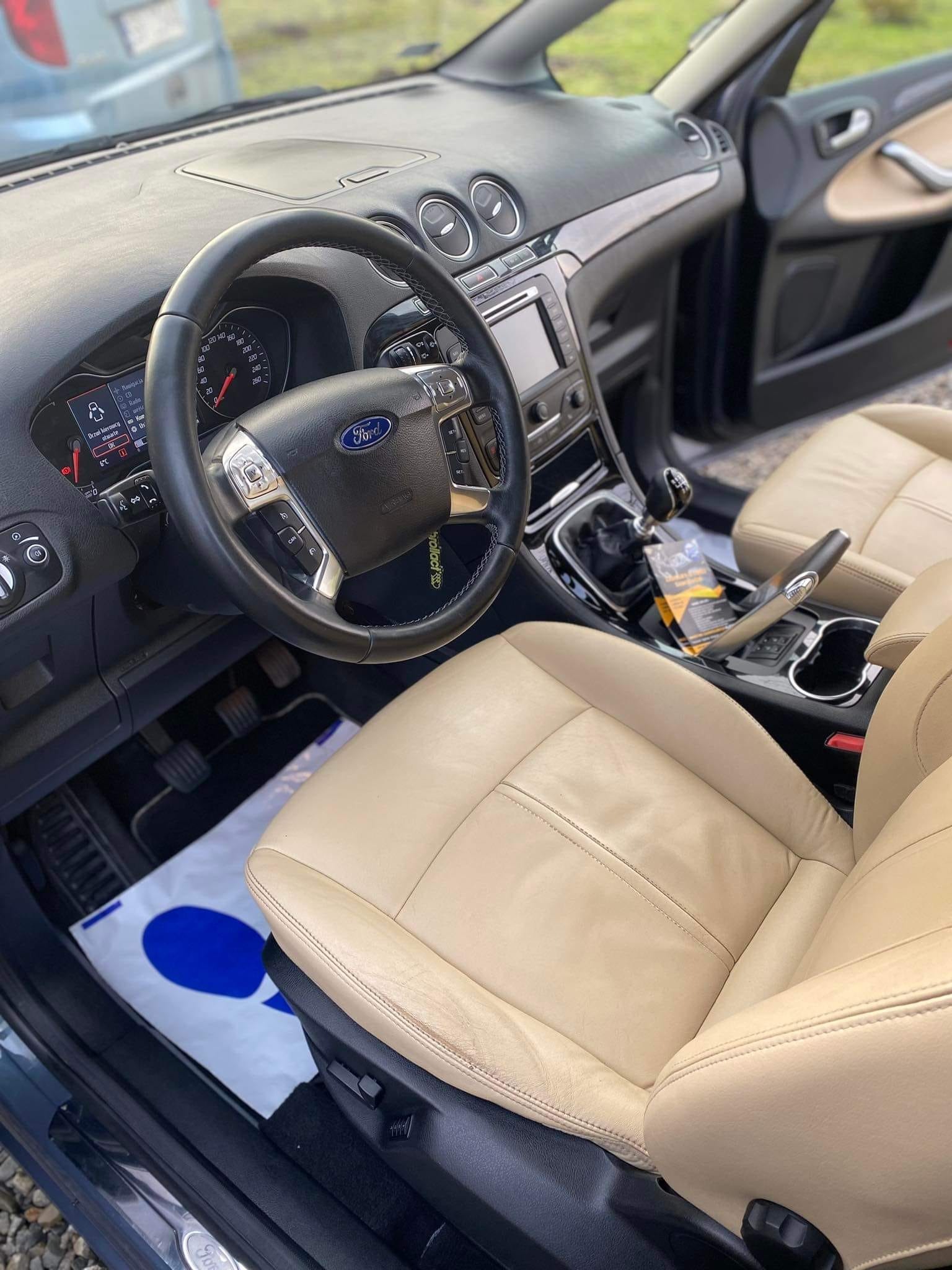 Wnętrze samochodu Ford S-Max z beżową skórzaną tapicerką foteli, widok z perspektywy kierowcy, na podłodze papierowa mata ochronna z niebieskim nadrukiem.