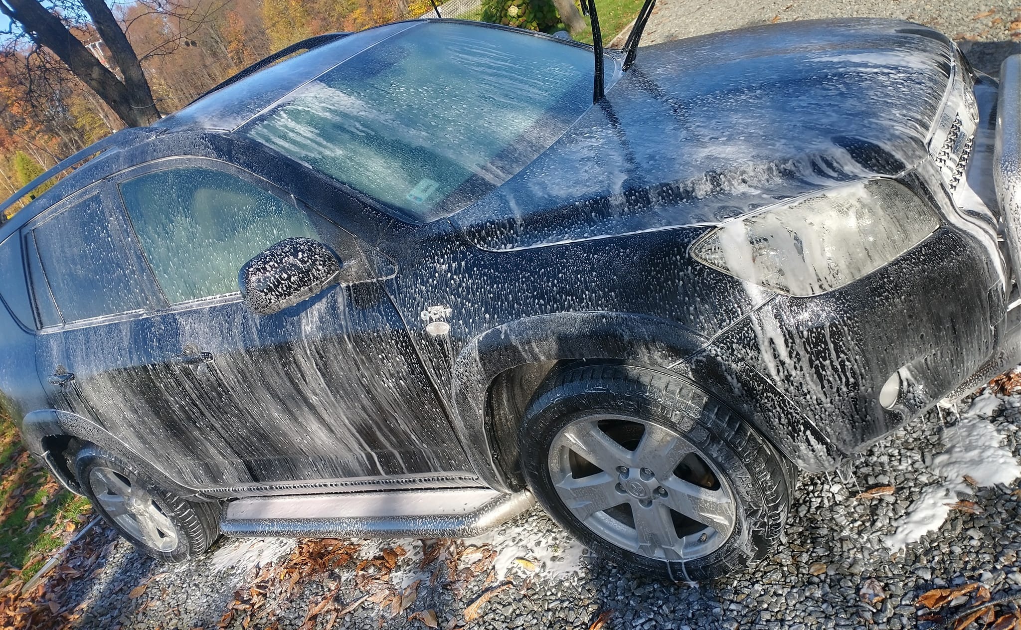 Czarny SUV pokryty pianą podczas mycia na zewnątrz, widoczne krople wody spływające po karoserii, jesienne liście na ziemi.