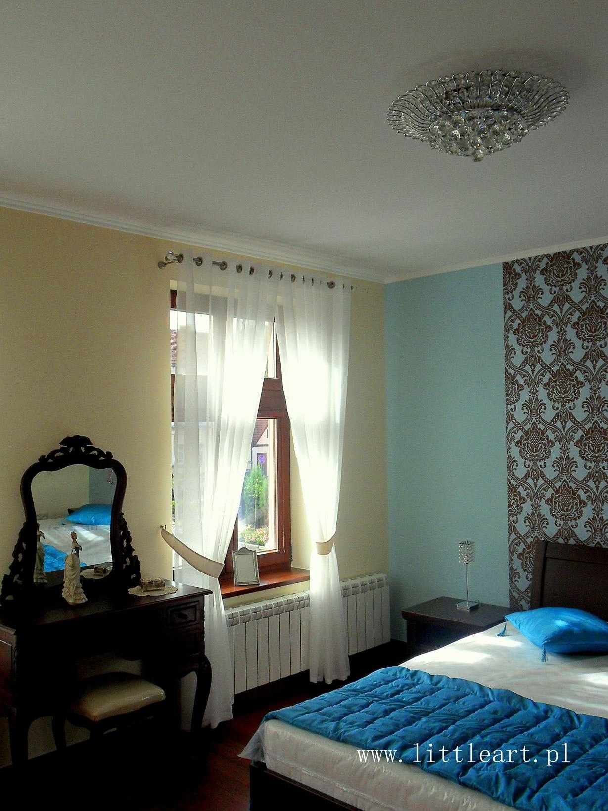 Jasna sypialnia z oknem przysłoniętym zwiewnymi, białymi zasłonami, toaletka z rzeźbionym lustrem i statuetką, oraz łóżko z niebieską narzutą, akcentujące klasyczny wystrój.