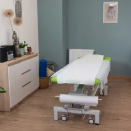 Gabinet z leżanką do rehabilitacji, na ścianie grafika układu kostnego, obok szafka z kwiatami i sprzętem audio.