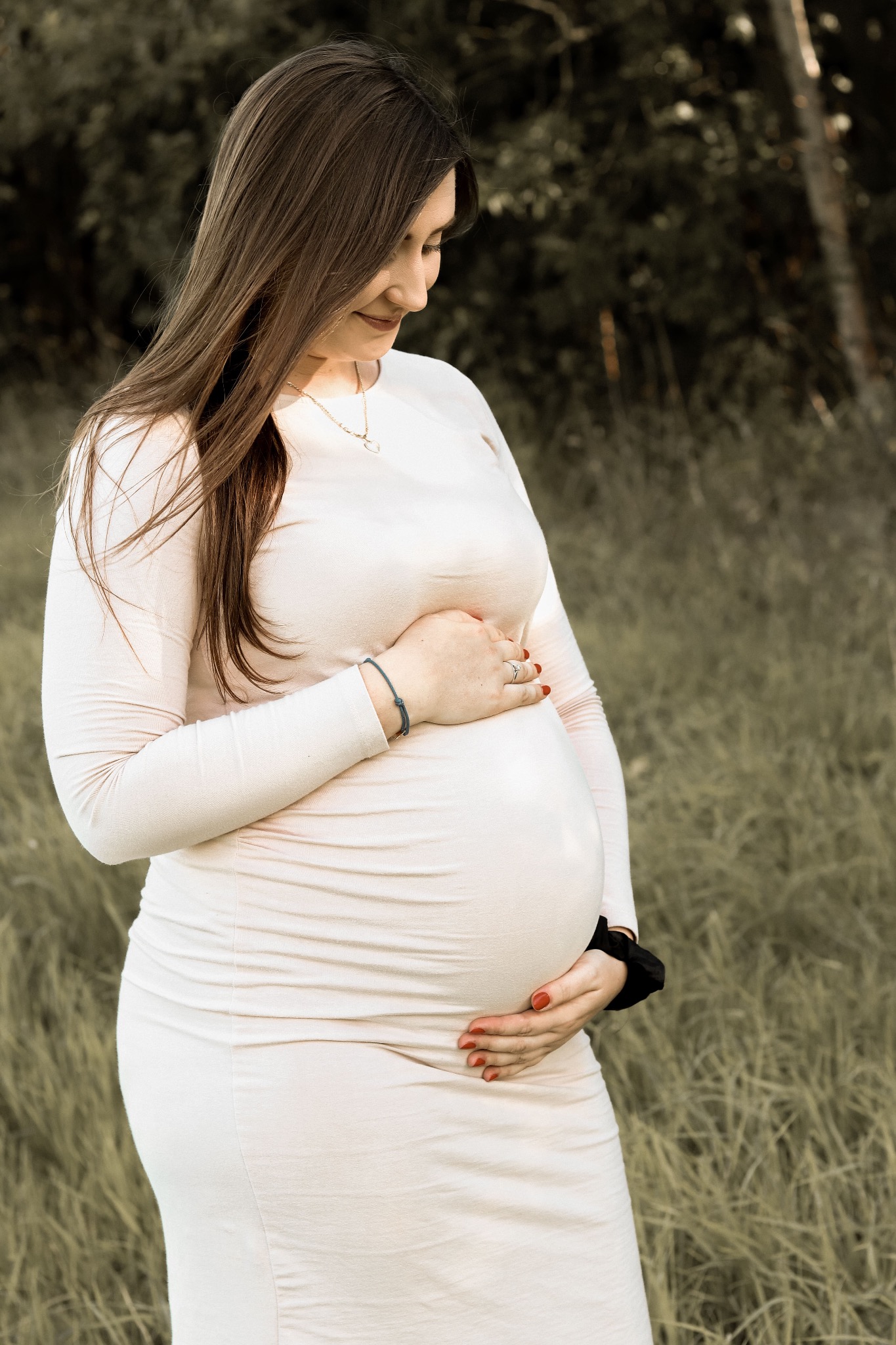 Kobieta w jasnej sukience ciążowej delikatnie obejmuje brzuch, spoglądając w dół, na tle jesiennej trawy.