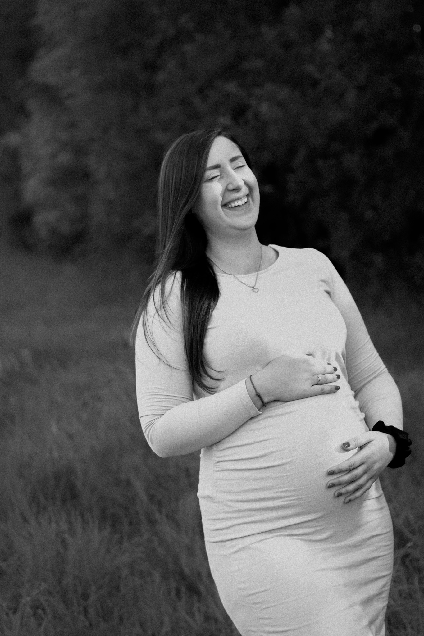 Czarno-białe zdjęcie uśmiechniętej kobiety w dopasowanej sukience, trzymającej ręce na brzuchu, w tle rozmyte drzewa i trawa.