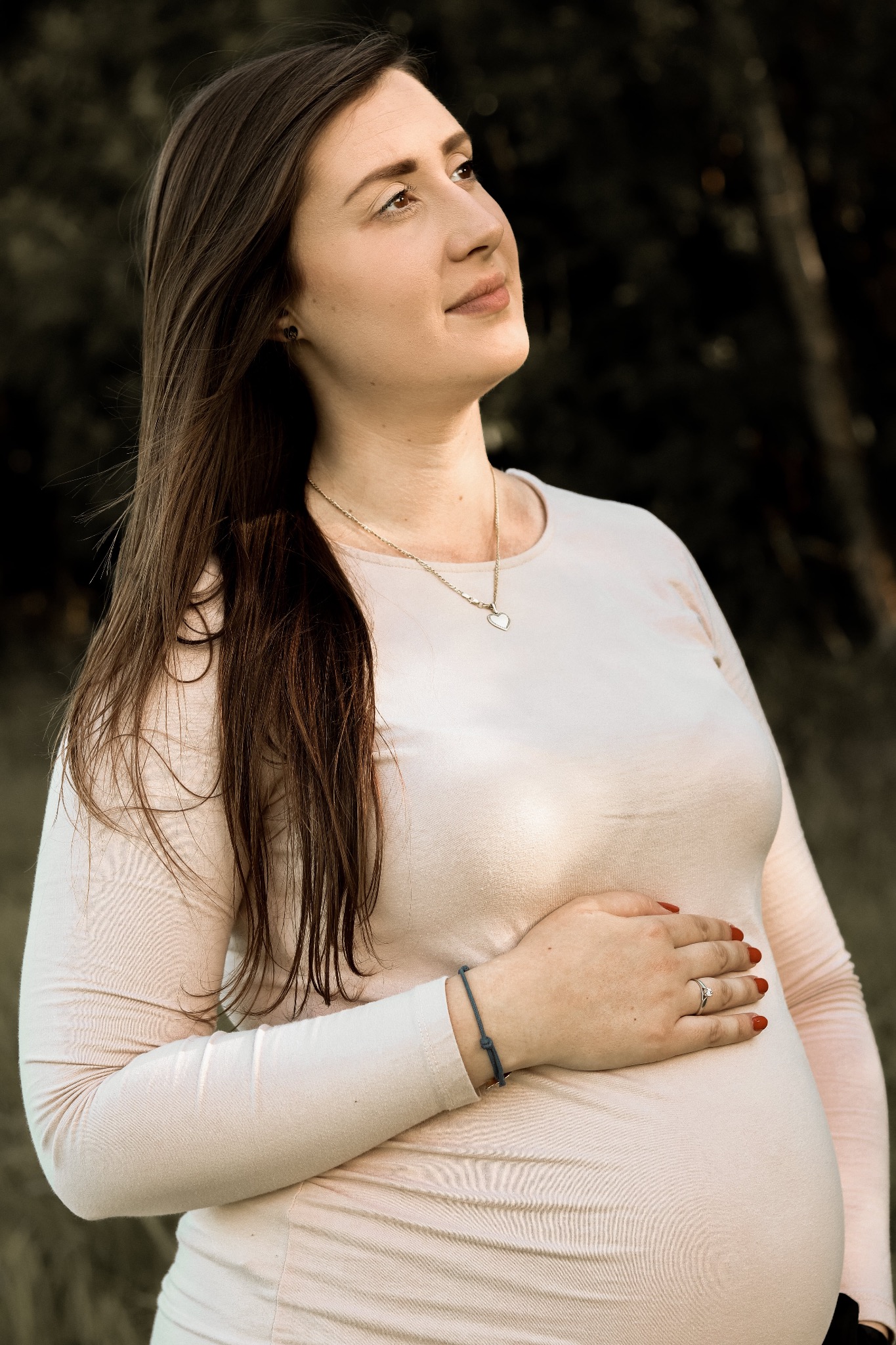 Portret kobiety w ciąży, z ręką na brzuchu, spoglądającej w górę, w tle zieleń.