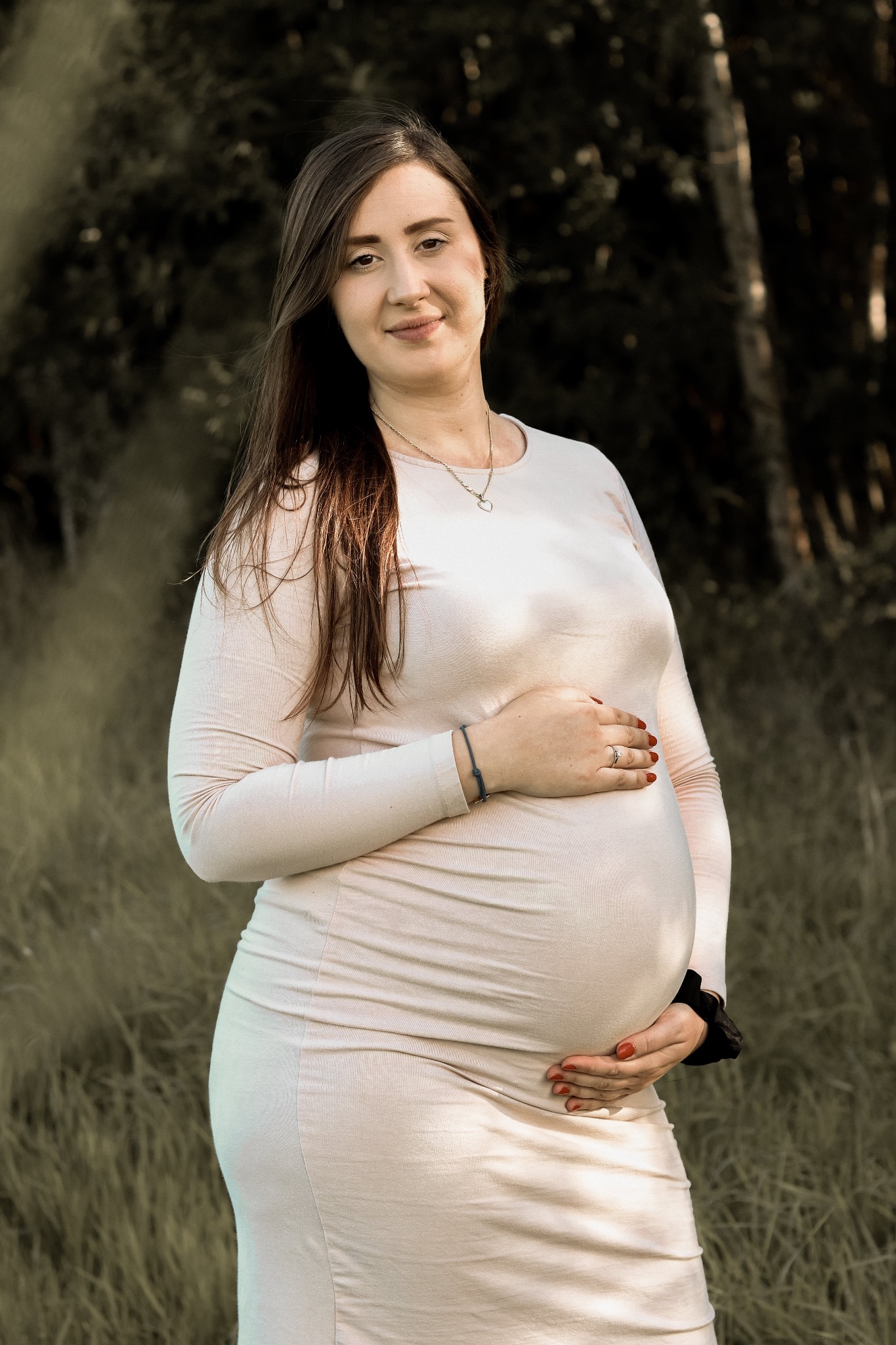 Uśmiechnięta, przyszła mama w dopasowanej, jasnoróżowej sukience, trzyma dłonie na brzuchu, pozując na tle leśnej scenerii.
