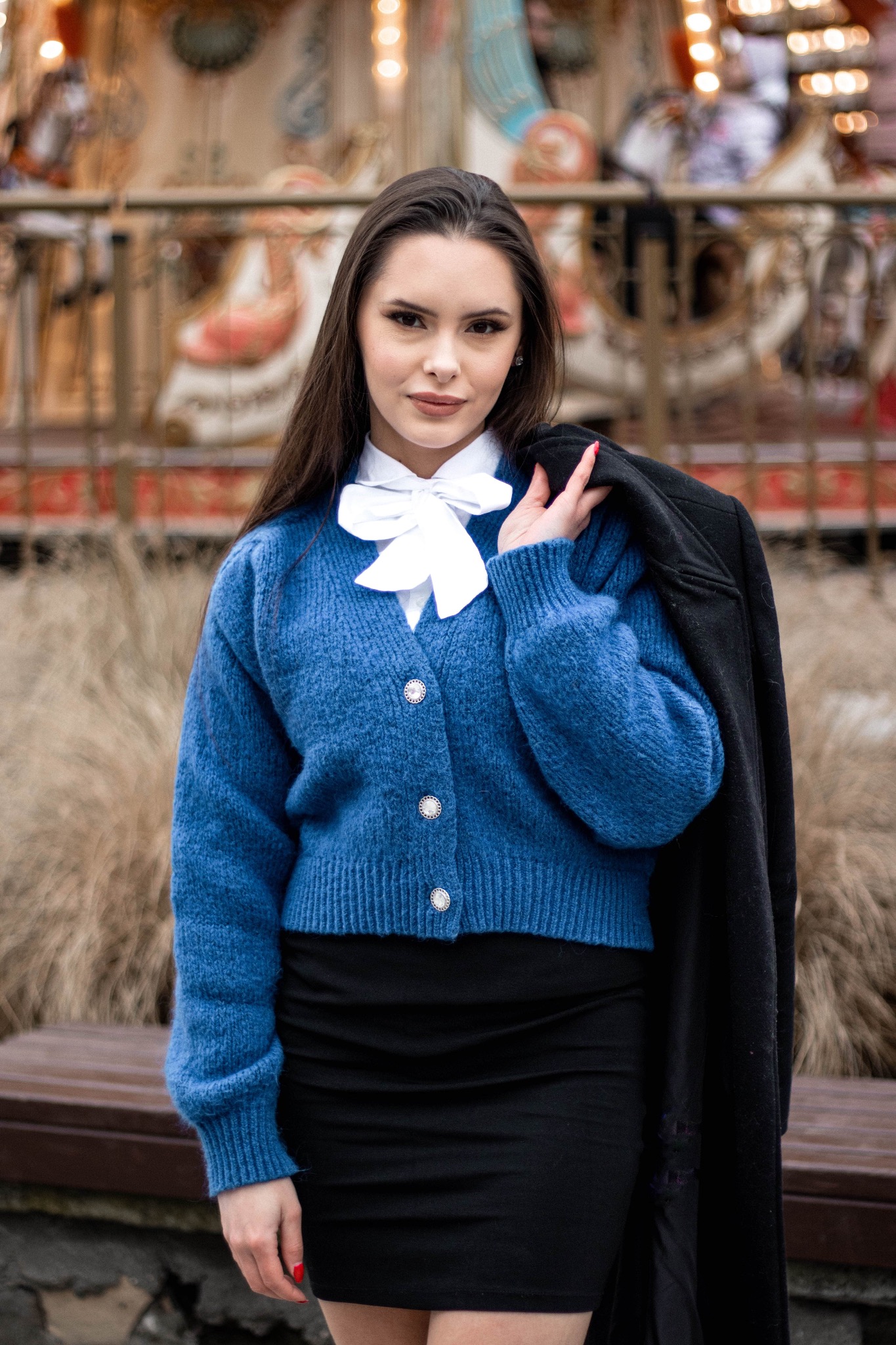 Portret kobiety w niebieskim kardiganie z białą kokardą, trzymającej czarny płaszcz na ramieniu, na tle karuzeli.