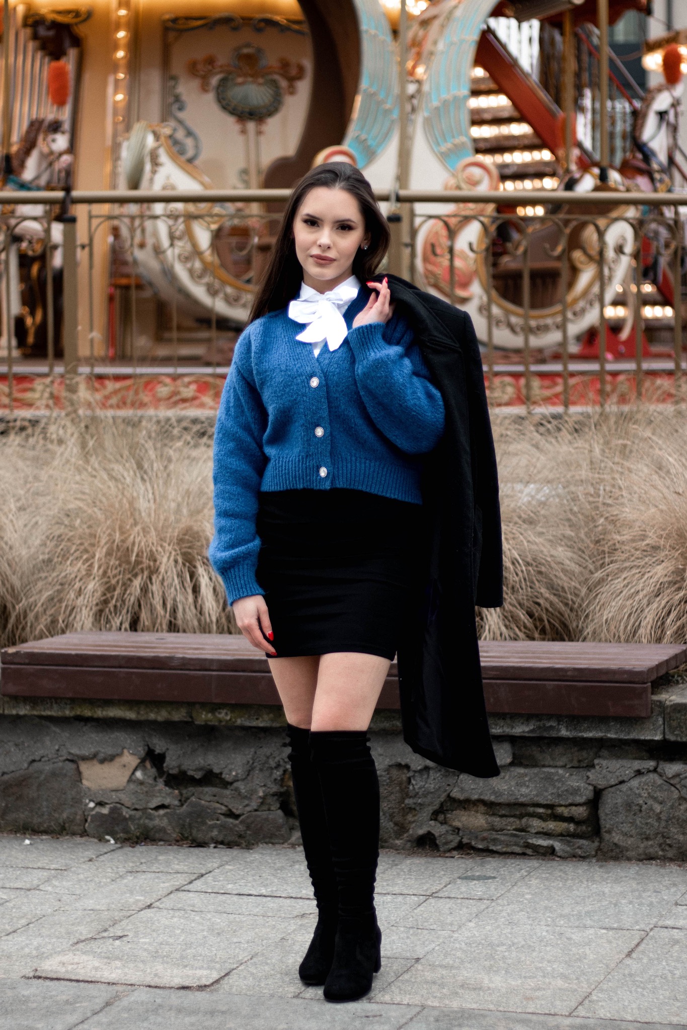 Portret kobiety w niebieskim swetrze i czarnej spódnicy, z marynarką na ramieniu, na tle karuzeli i ławki.