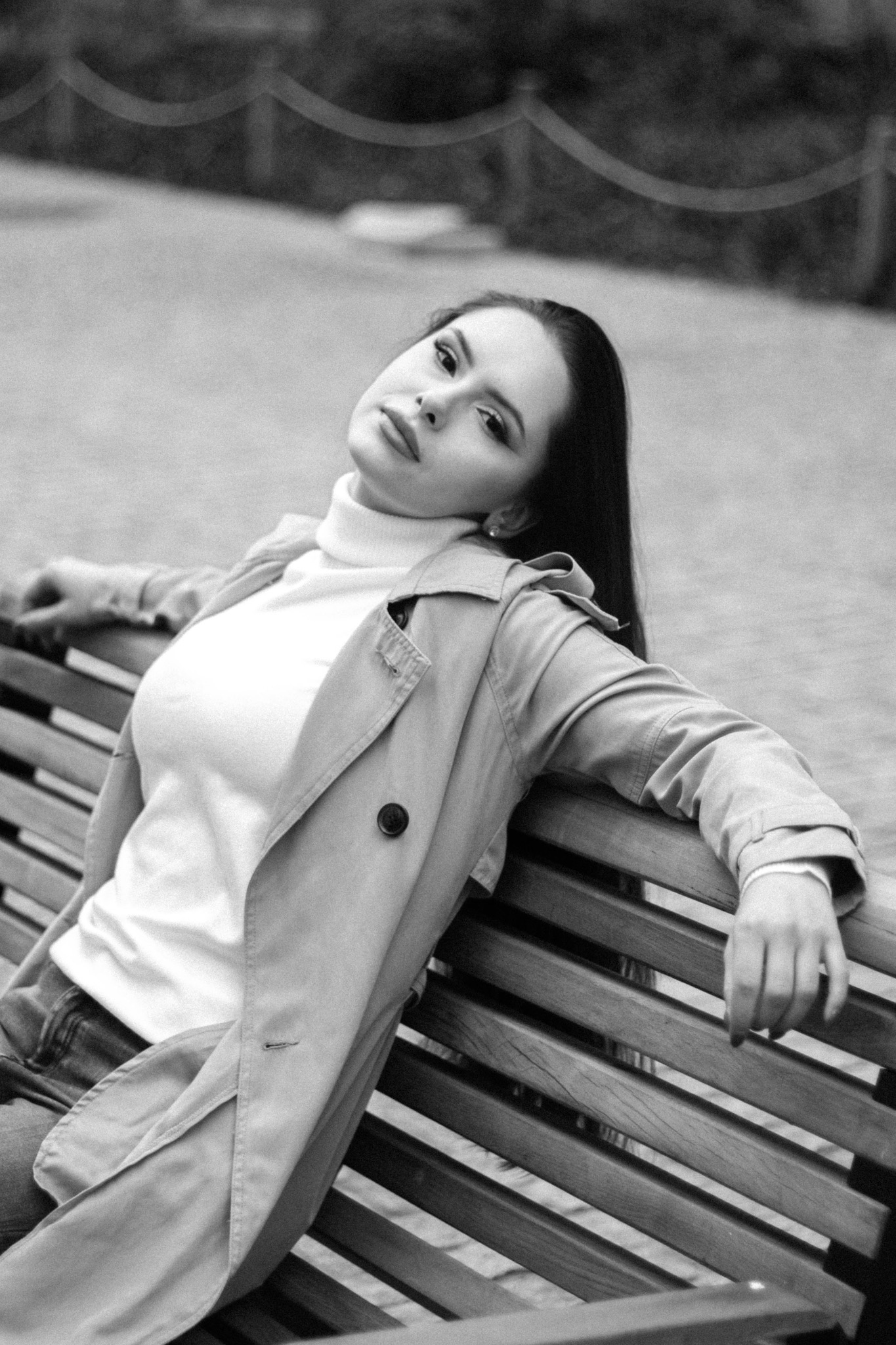 Portret kobiety w jasnym płaszczu i golfie, oparta o drewnianą ławkę na tle parku, czarno-biała fotografia.