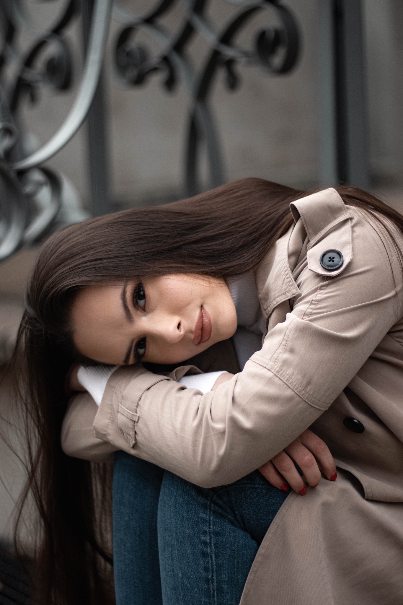 Portret kobiety w beżowym płaszczu, opierającej głowę na rękach, z widocznym fragmentem metalowej balustrady w tle.
