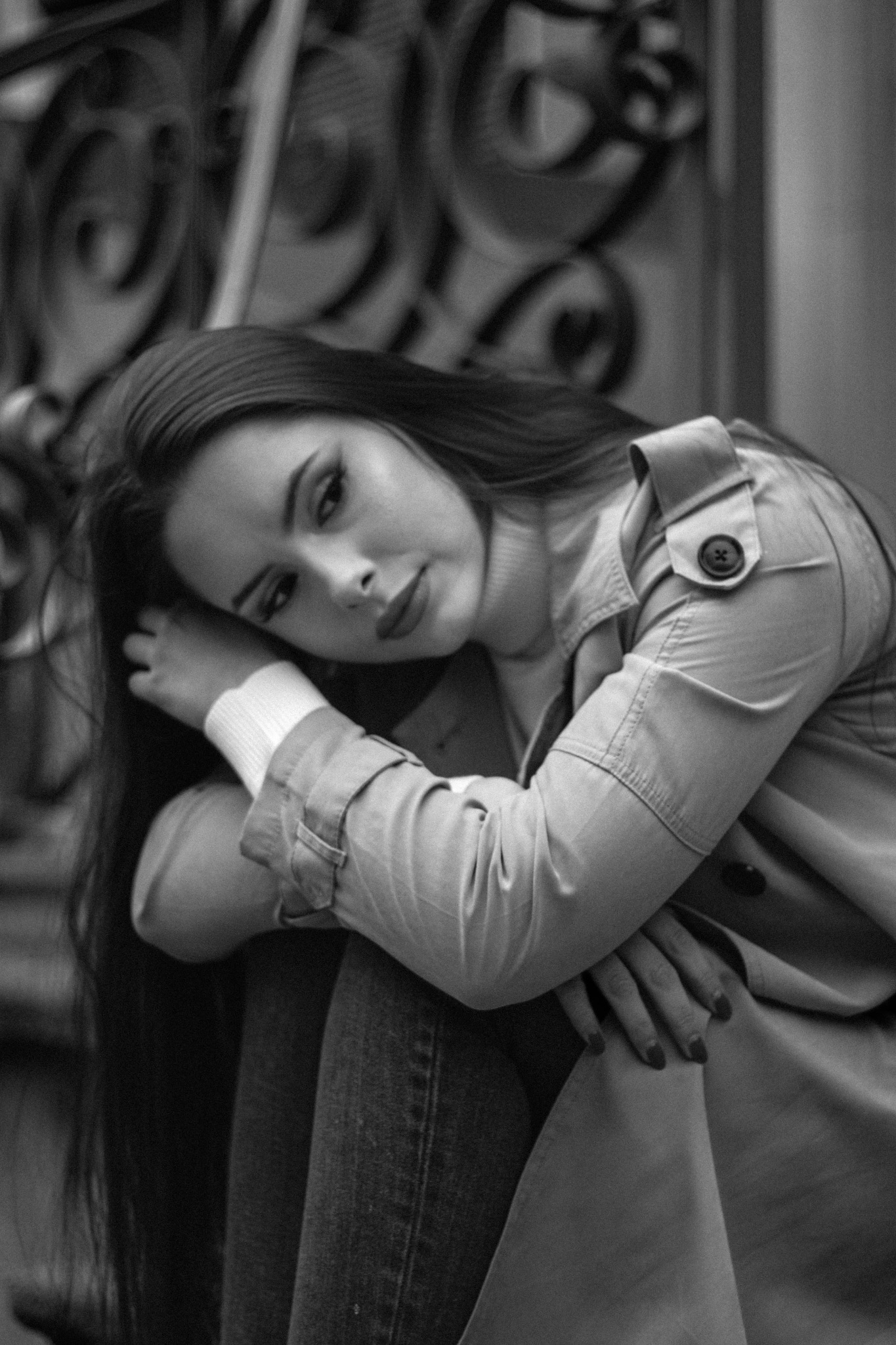 Portret kobiety w płaszczu, opierającej głowę na dłoni, w czarno-białej tonacji, z dekoracyjnym metalowym tłem.