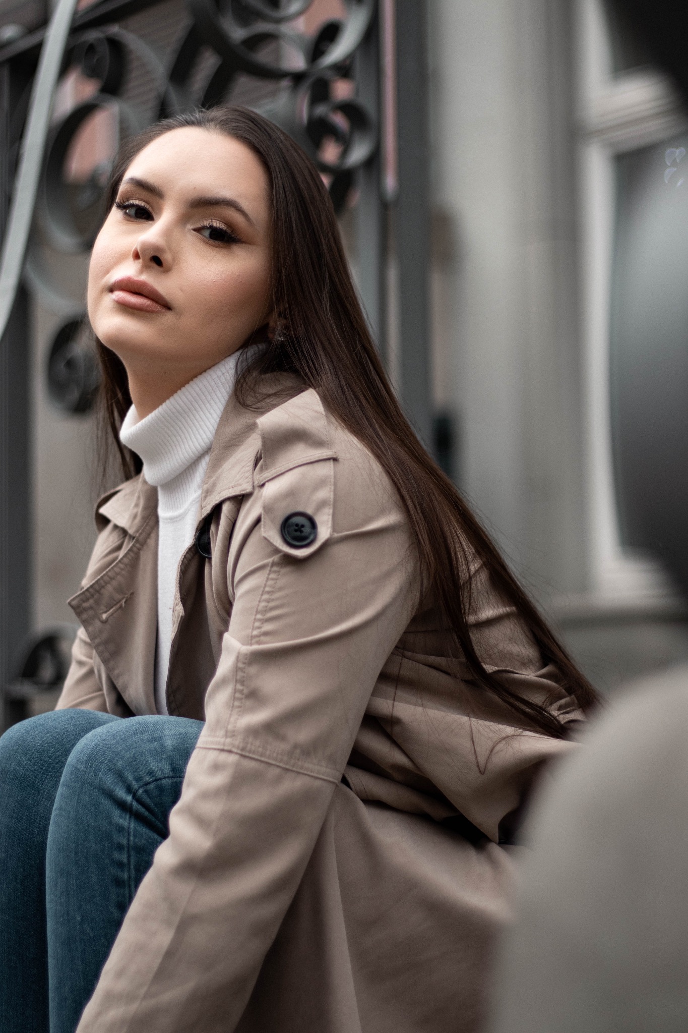 Portret kobiety w beżowym trenczu i białym golfie, siedzącej na schodach z metalową balustradą, z głową lekko odchyloną do tyłu.