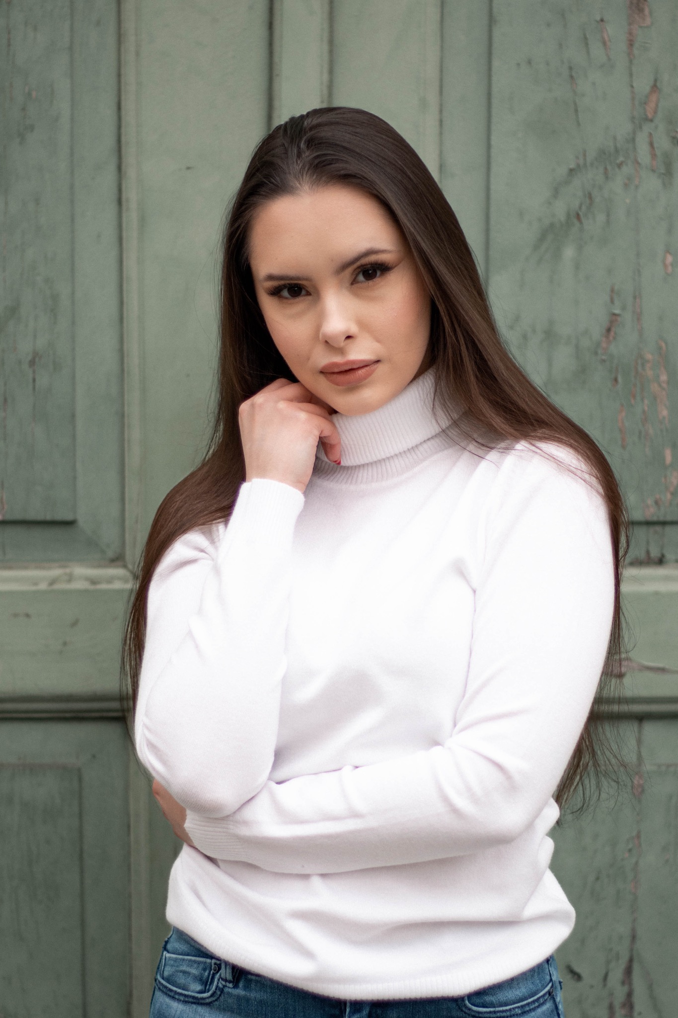 Portret kobiety w białym golfie na tle zielonych, lekko zniszczonych drzwi.