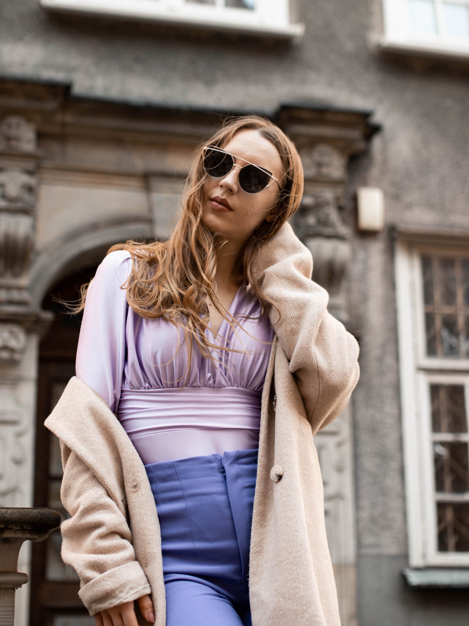 Portret kobiety w okularach przeciwsłonecznych, w pastelowej bluzce i płaszczu, pozującej na tle architektonicznej fasady budynku.