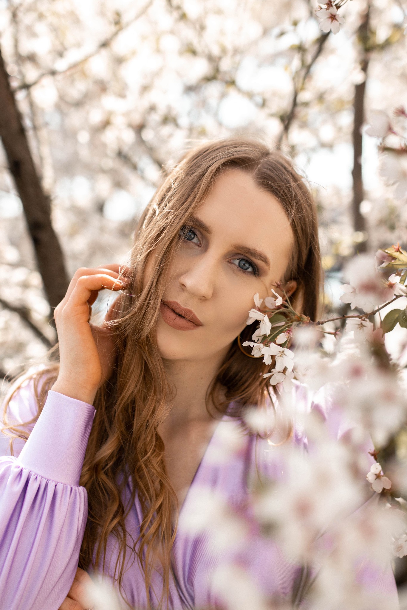 Portret kobiety o blond włosach i niebieskich oczach w liliowej sukience, pozującej na tle kwitnących drzew z białymi kwiatami.