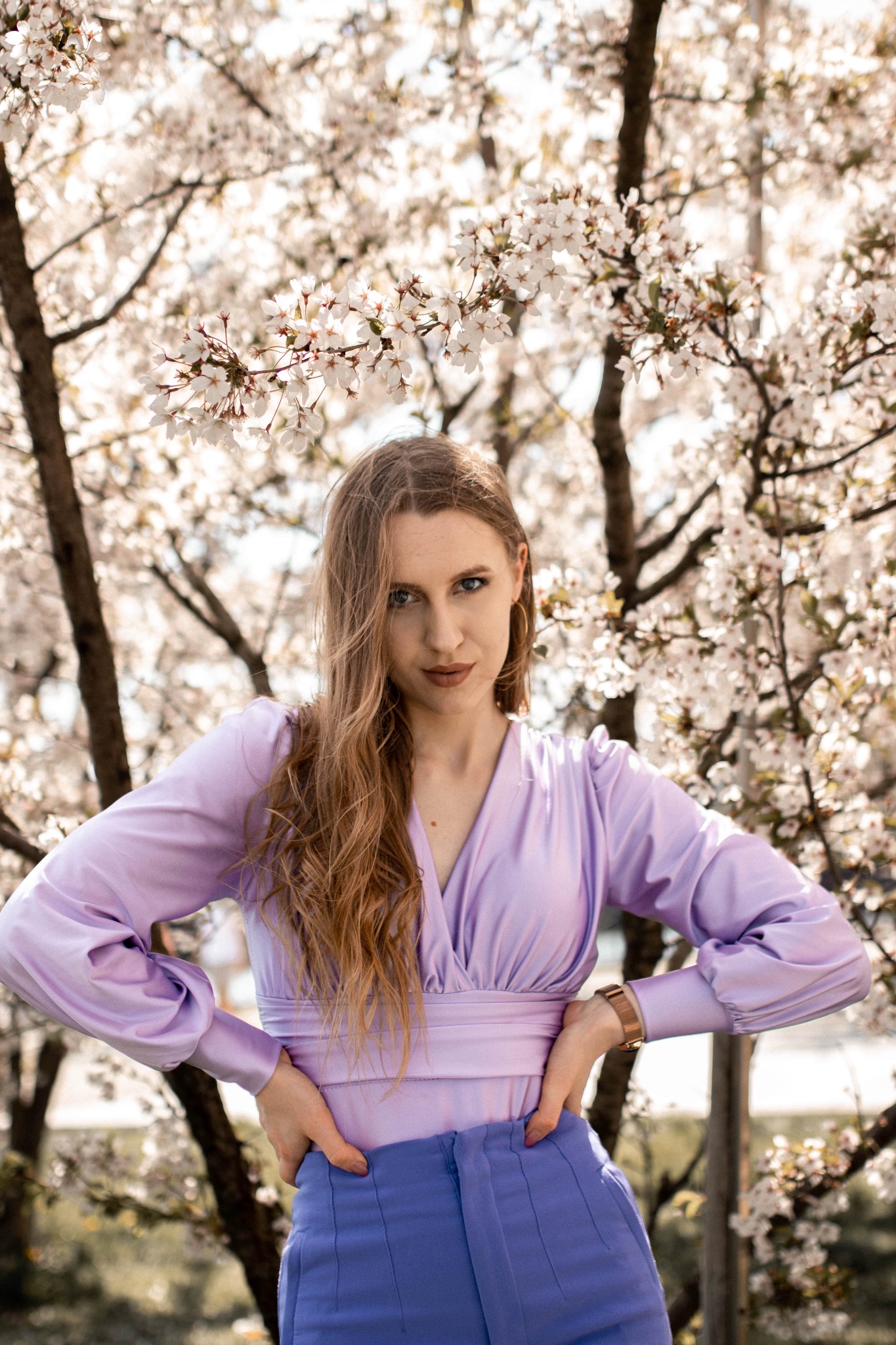 Kobieta w liliowym topie i niebieskich spodniach pozuje na tle kwitnącego drzewa z białymi kwiatami.
