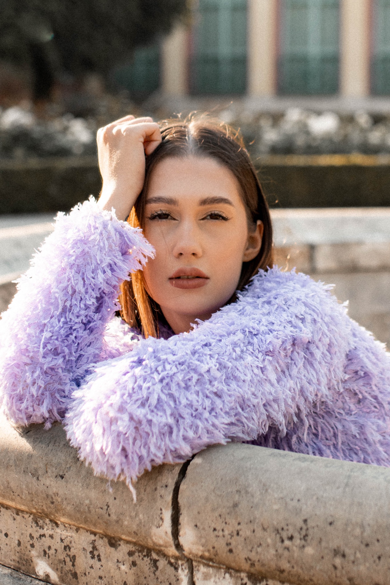 Portret młodej kobiety w puszystej, liliowej kurtce, opierającej się o kamienną balustradę w plenerze.