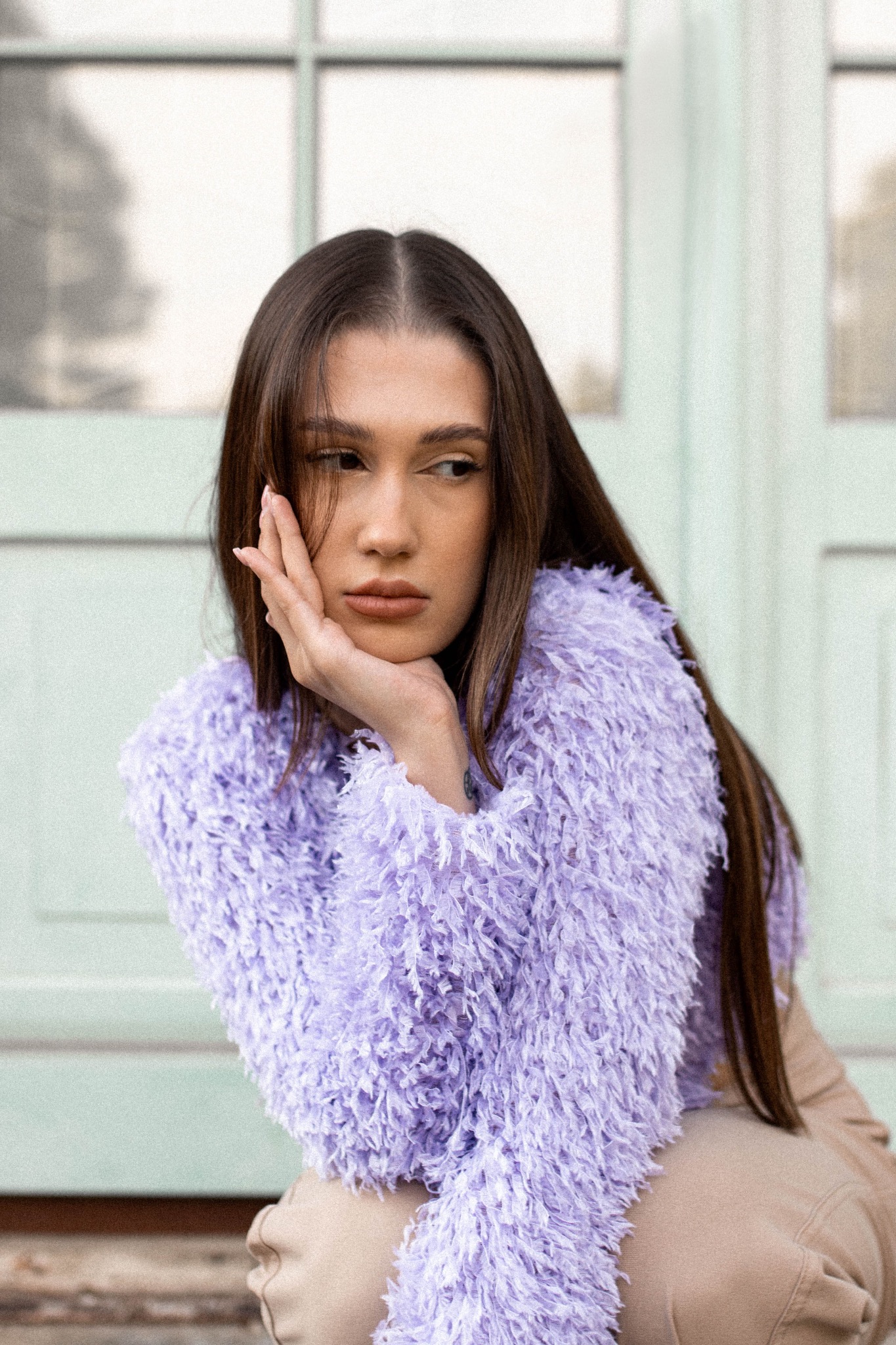 Portret młodej kobiety w puszystym, liliowym swetrze, opierającej policzek na dłoni, zamyślony wzrok skierowany w bok, na tle jasnych, przeszklonych drzwi.