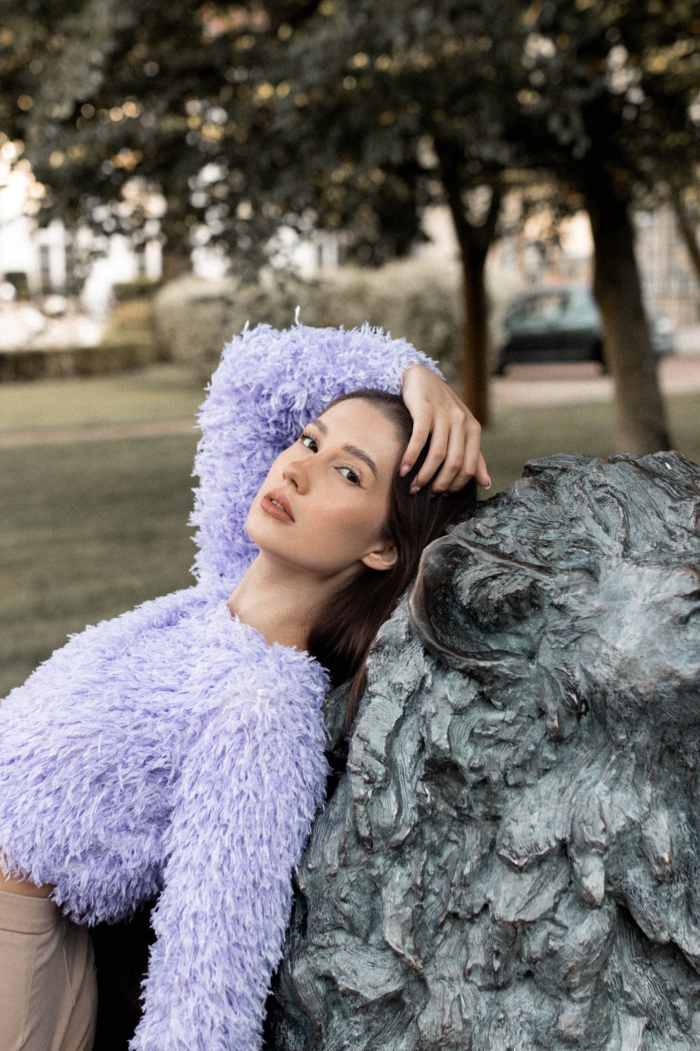 Portret kobiety w liliowym swetrze, opierającej się o kamienną rzeźbę w parku, z delikatnym makijażem i naturalnym oświetleniem.