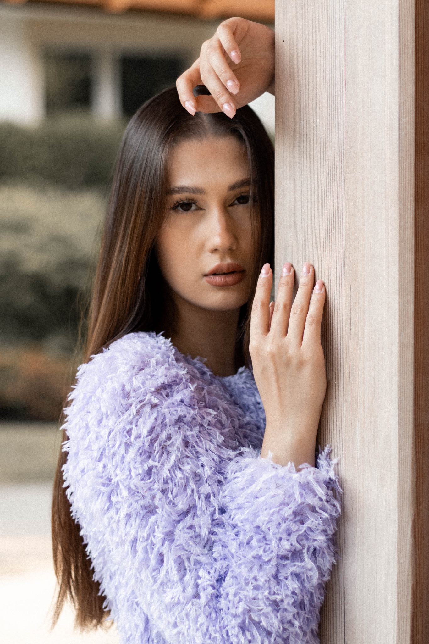 Portret młodej kobiety opierającej się o drewnianą ścianę, ubrana w futrzany, liliowy sweter, z dłońmi delikatnie dotykającymi ściany.