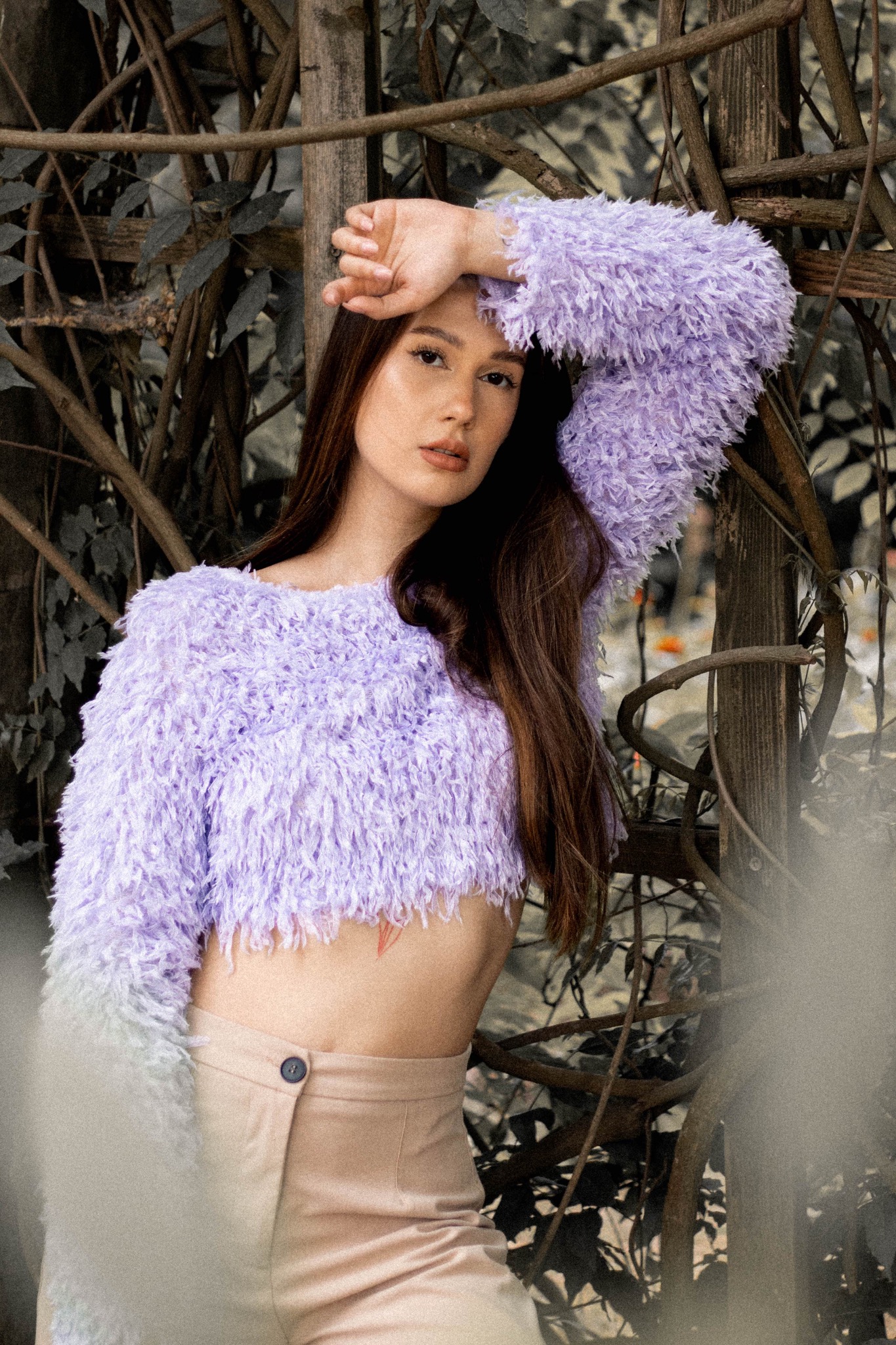Portret kobiety w fioletowym, futrzanym swetrze i beżowych spodniach, pozującej na tle gałęzi i liści.