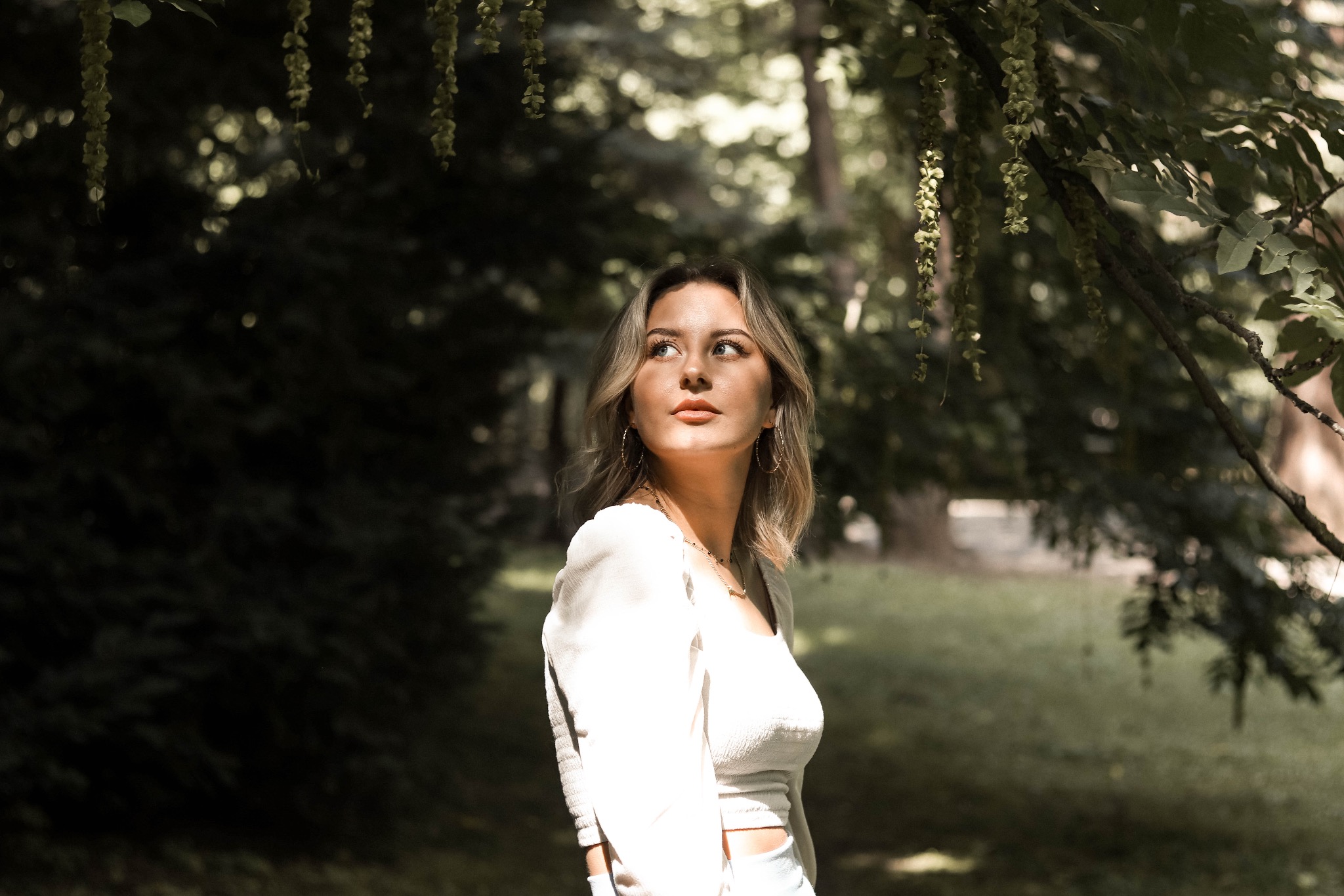 Portret kobiety w białej bluzce, spoglądającej w lewo, na tle zieleni drzew w parku, z wiszącymi kwiatostanami nad głową.