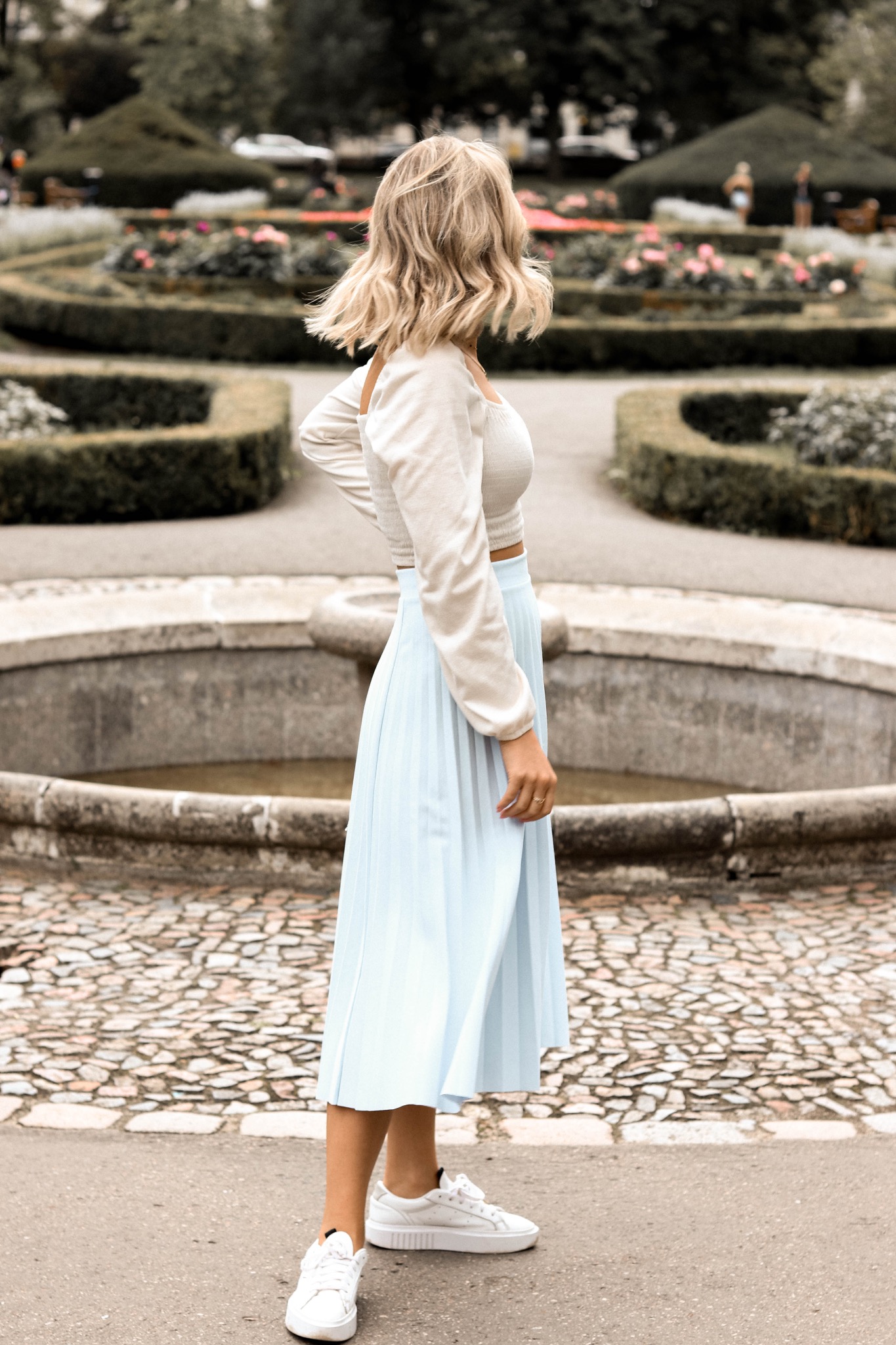 Kobieta w jasnej stylizacji, stojąca tyłem na tle fontanny i zadbanego parku z geometrycznymi rabatami.