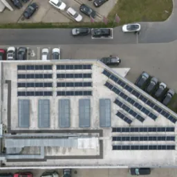 Widok z drona na dach budynku z panelami słonecznymi, systemem wentylacji i zaparkowanymi samochodami.