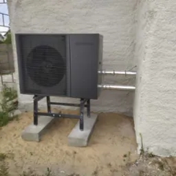 Czarna jednostka zewnętrzna z logo Samsung, zamontowana na metalowej konstrukcji na betonowych podstawach, z podłączonymi rurami do ściany budynku o szorstkiej fakturze.