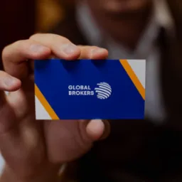 Dłoń mężczyzny trzyma wizytówkę firmy Global Brokers z logo w kolorach niebieskim, białym i żółtym.