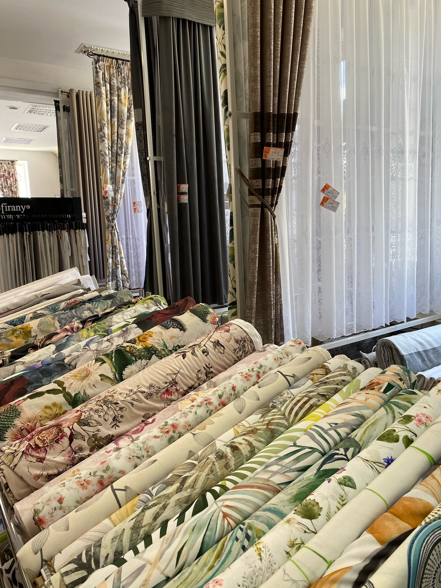 Szeroki wybór tkanin w rolkach z motywami kwiatowymi i roślinnymi, prezentowany na tle gotowych zasłon w sklepie z tekstyliami.