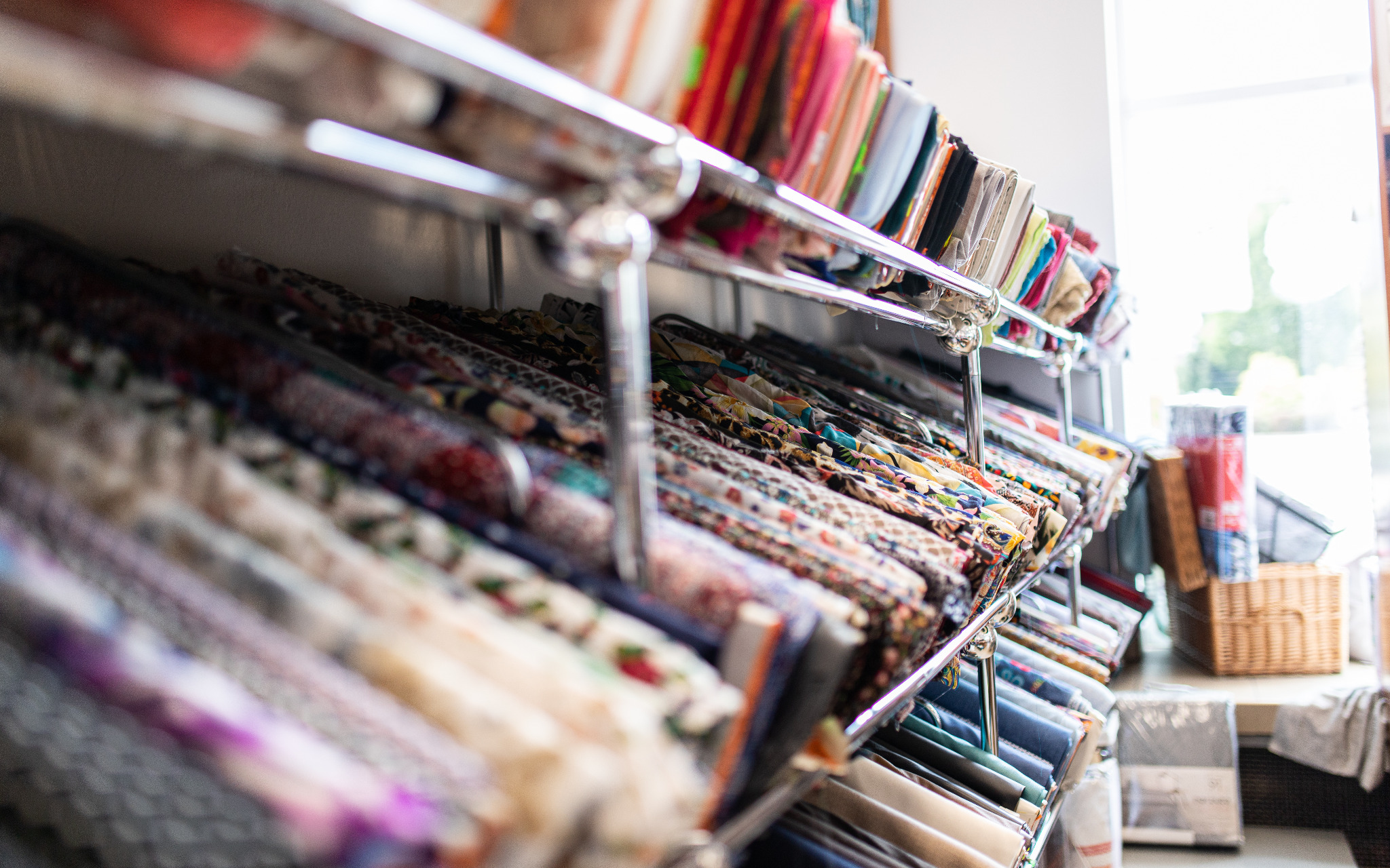 Regał z różnorodnymi, kolorowymi tkaninami w hurtowni; widoczne wzory kwiatowe, geometryczne i abstrakcyjne, ułożone równo na metalowych półkach, z oknem w tle.