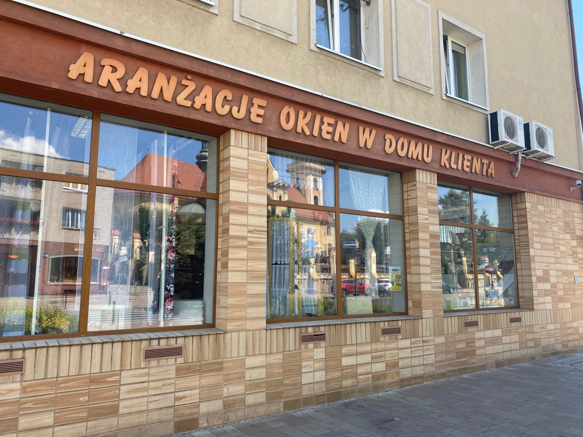 Fasada budynku z napisem 'Aranżacje okien w domu klienta' nad witrynami, w których widać firany i zasłony, odbicie kościoła w szybach.