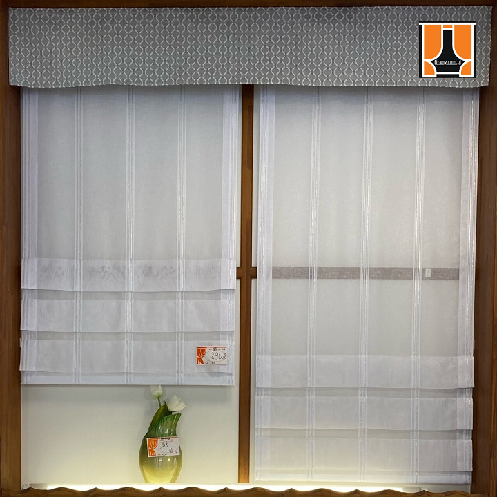 Dwie rolety rzymskie z delikatnego, białego materiału w drewnianej ramie okiennej, z dekoracyjnym szarym panelem u góry i wazonem z białymi tulipanami u dołu po lewej stronie.