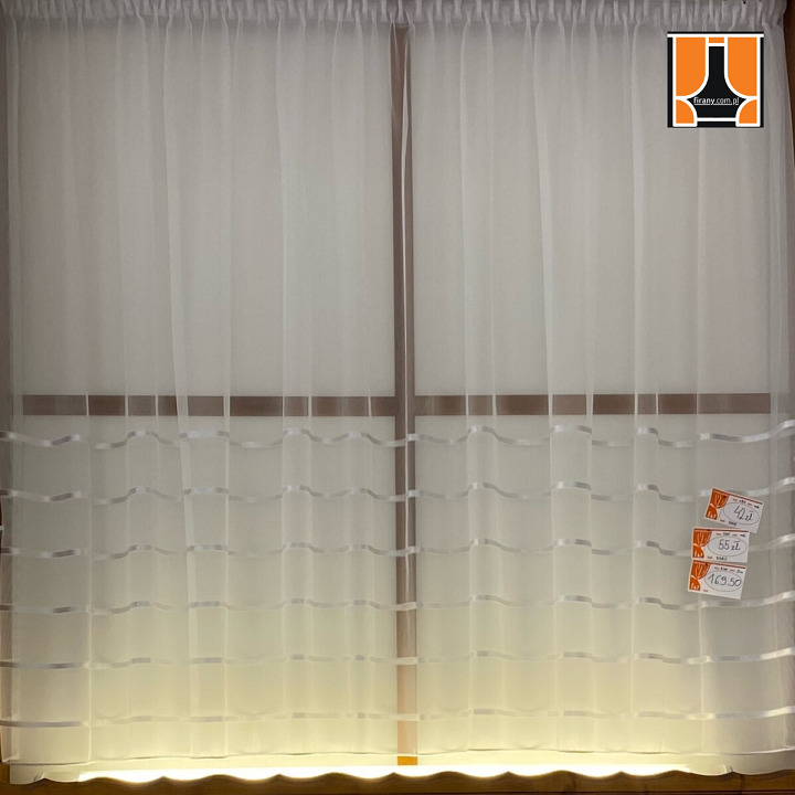Delikatna, biała firanka z poziomymi, falistymi wstążkami na oknie, z widocznymi cenami na etykietach i logo firmy w prawym górnym rogu.