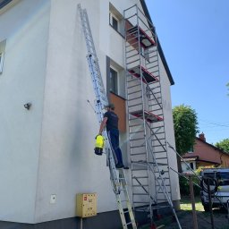 4Invest - Pracownik na drabinie z opryskiwaczem, przygotowujący się do mycia elewacji dwupiętrowego domu z rusztowaniem, widoczna skrzynka gazowa.