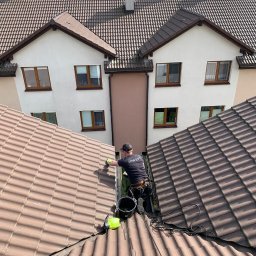 4Invest - Pracownik w uprzęży, z góry, czyści rynnę na dachu dwuspadowego domu z brązową dachówką, używając szczotki i wiadra, widoczny spryskiwacz.