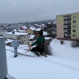 4Invest - Pracownik w uprzęży na zaśnieżonym dachu odśnieża go pomarańczową łopatą, w tle zabudowa miejska i pochmurne niebo.