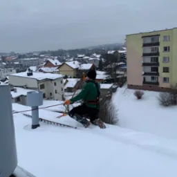 Pracownik w uprzęży na zaśnieżonym dachu odśnieża go pomarańczową łopatą, w tle zabudowa miejska i pochmurne niebo.