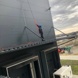 4Invest - Pracownik w niebieskim kasku czyści ciemnoszarą elewację hali przemysłowej za pomocą szczotki na długim teleskopowym kiju, stojąc na dachu; w tle widoczny podnośnik koszowy i generator prądu.