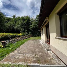 4Invest - Częściowo wyczyszczony, mokry chodnik z kostki brukowej przy elewacji domu, z widocznym odrostem chwastów w spoinach, ogród z zielenią w tle i żółty rowerek dziecięcy po lewej.