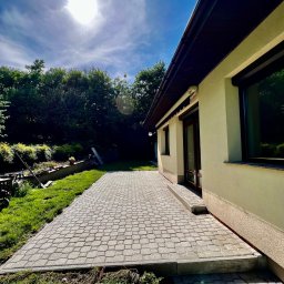 4Invest - Nowo ułożona, szara kostka brukowa tworzy patio przylegające do jasnego domu z ciemnymi oknami i dachem, widok na zielony trawnik i drzewa w tle, jasne słońce na niebie.